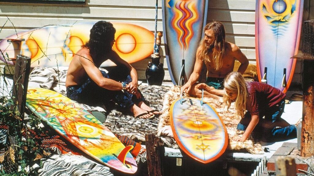 Imagen de surfistas en un entorno al aire libre, personalizando sus tablas de surf con llamativos diseños coloridos. Esta escena representa la cultura del surf y su estrecha conexión con el arte y la expresión personal.